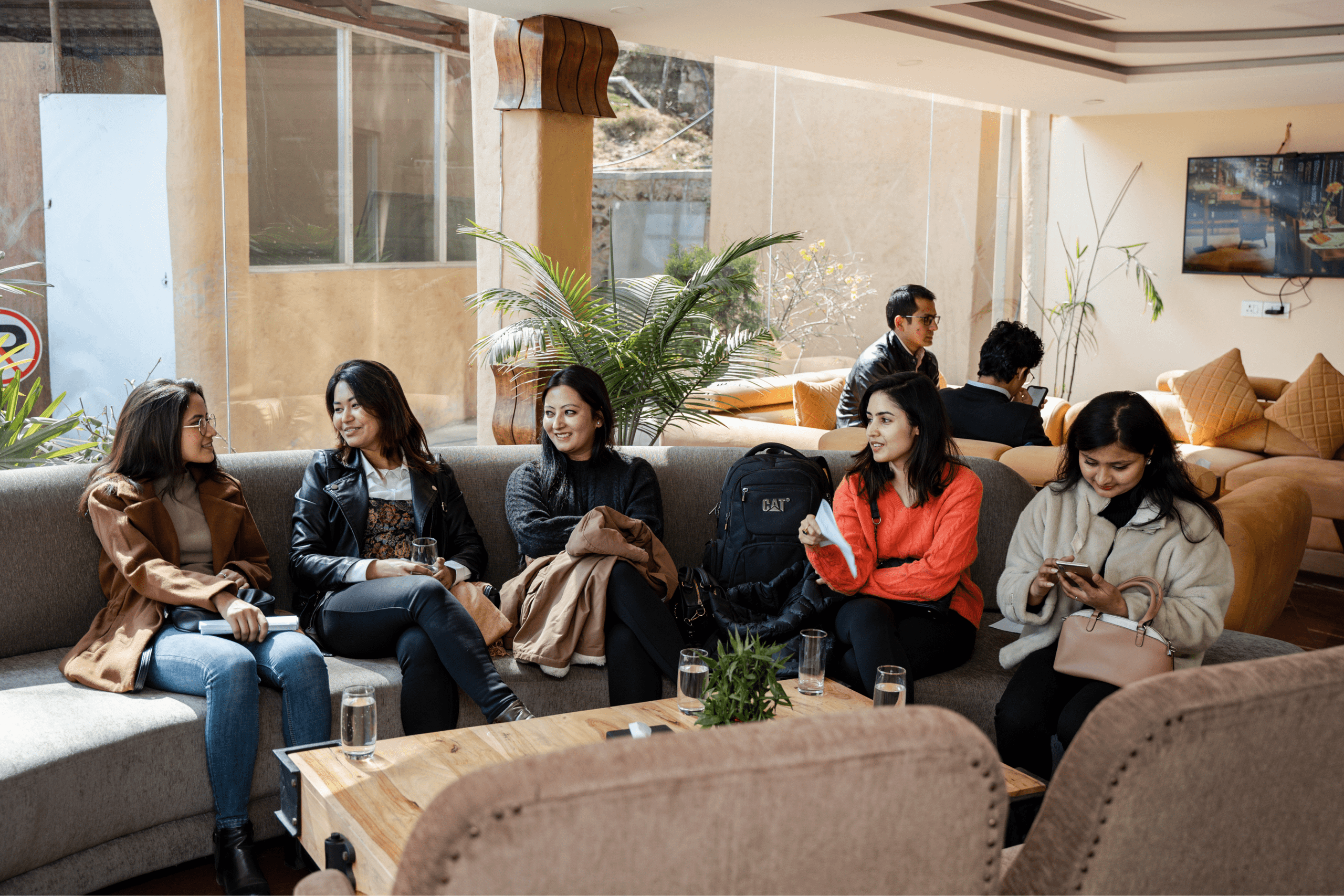 Four women sit on a couch, three of whom are listening to someone talk out of frame. They&#039;re dressed casually, two wear sweaters and one a leather jacket. They all have dark, straight or wavy hair.