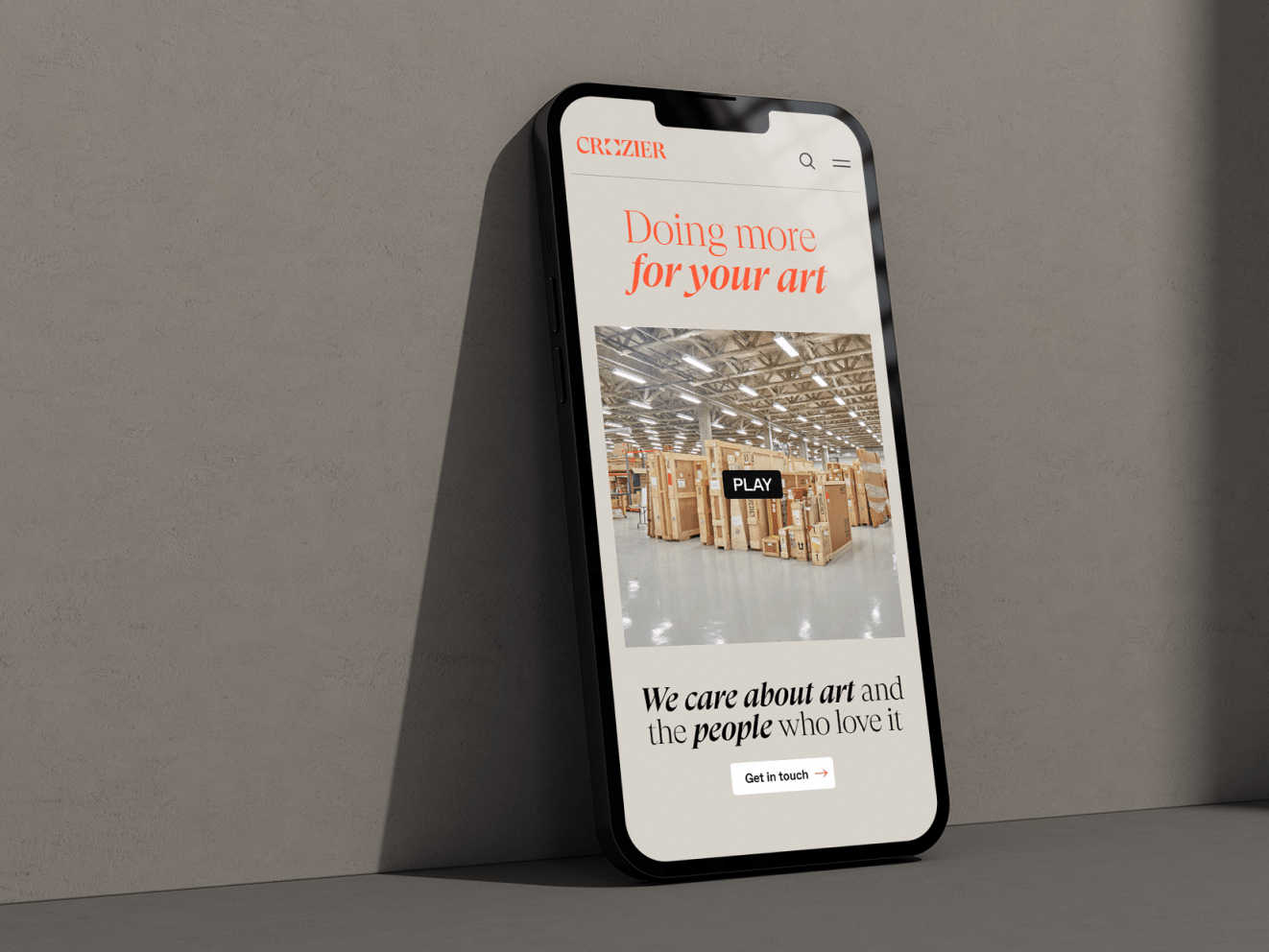 An iphone leaning against a grayish wall. On the screen is the Crozier homepage, featuring orange and black text that reads: "Doing more for your art" and "We care about art and the people who love it."