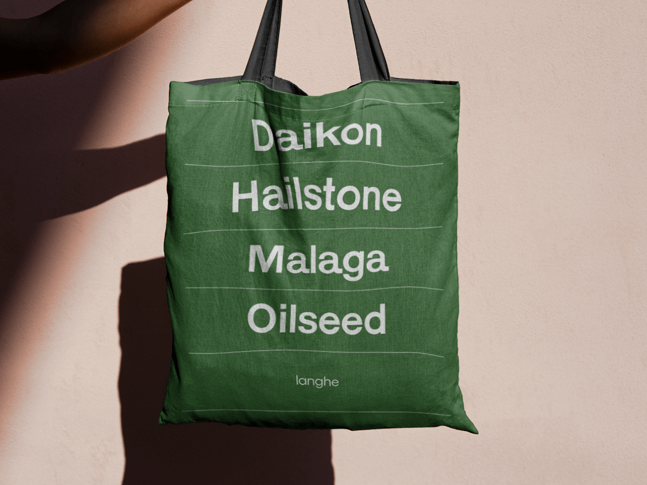 A forest green totebag being held against a tan wall. White text on the bag reads "Daikon, Hailstone, Malaga, Oilseed" and "Langhe."