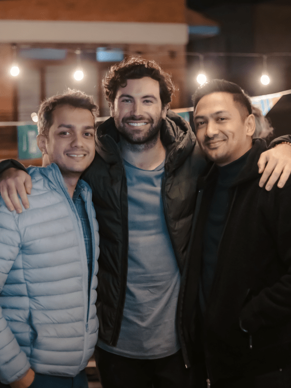 Outside's three founders, Atulya, Vincent, and Sujan, smiling. Vincent has his arms wrapped around the other two.