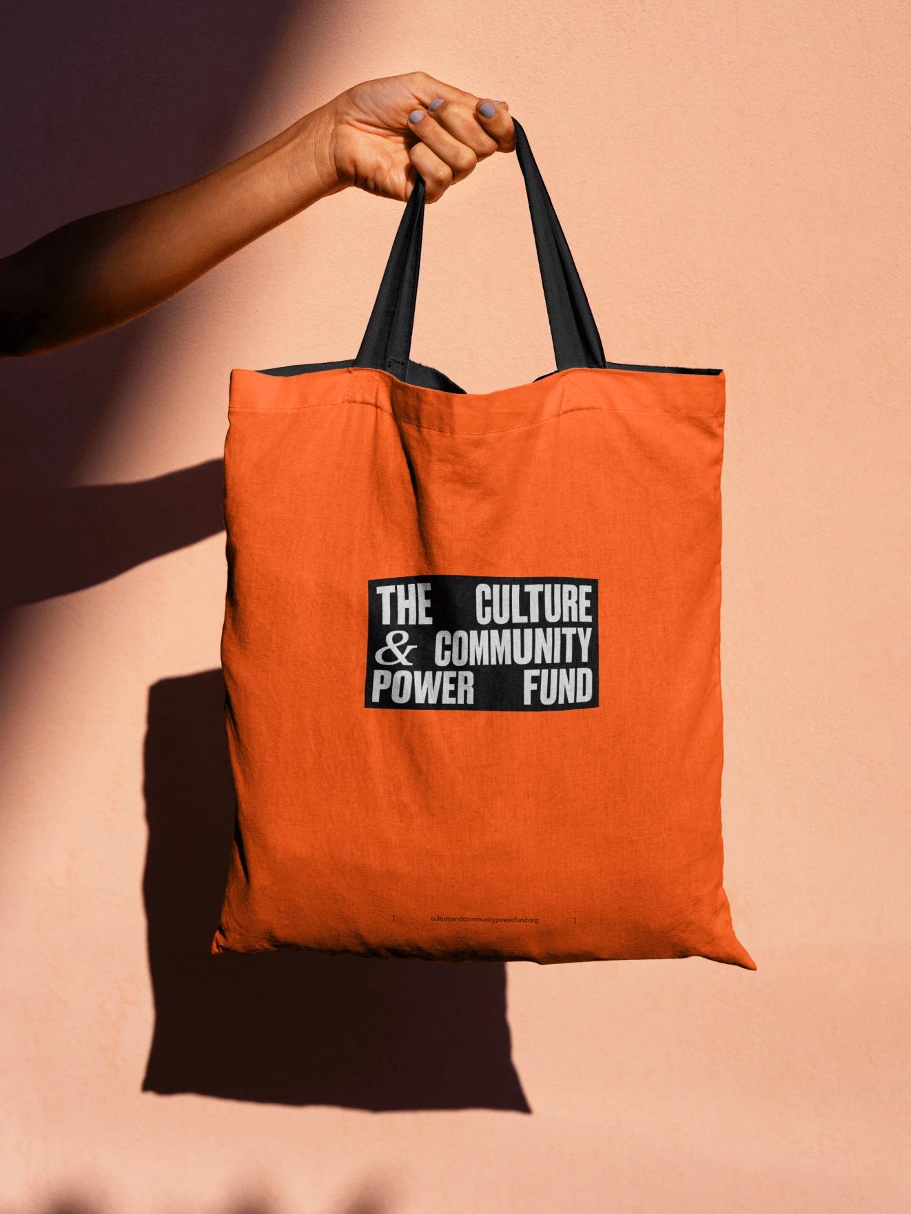 The Culture &amp; Community Power Fund logo on white text in a black rectangle, on a bright orange tote bag.